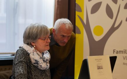 Een oudere vrouw zit aan een computer. Een man met wit haar kijkt mee over haar schouder.