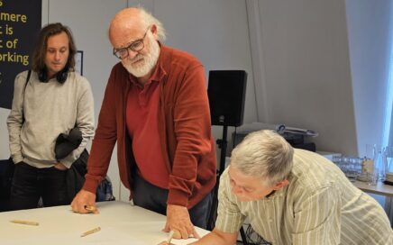 Een oudere man en vrouw noteren op een groot blad. Een jongere man kijkt mee over de schouder.
