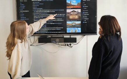 Histories medewerkers Morgane en Sajara kijken naar een scherm. Een Wikipediapagina van Brussel staat open. Morgane wijst en Sajara kijkt.