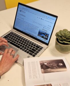 De Wikipediapagina van Geraardsbergen staat open op een computer. Je ziet twee handen met zilveren ringen, een cactus en een boek dat open ligt.