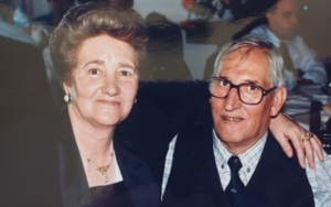 Een oudere vrouw en man staan glimlachend op de foto. De vrouw heeft haar arm rond hem.