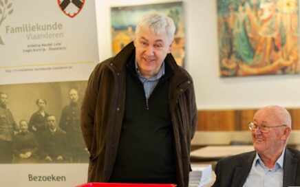 Twee oudere mannen lachen naar de camera. Eén man zit met een boek in zijn hand. De andere staat recht.