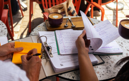 Twee mensen zitten op een terras met koffie. Er ligt een grote map open en een notitieboek. De linkerpersoon schrijft iets op een post-it.
