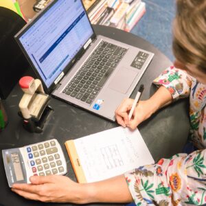 Een vrouw achter de computer. Ze is bezig op een rekenmachine en schrijft iets in een boekje.