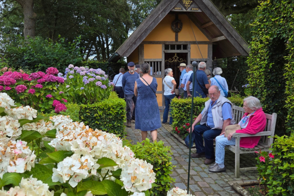 Mensen zitten op een bank, anderen kijken in een huis. Er staan bloemen op de voorgrond